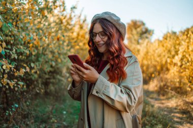 Neşeli gülümseyen ve cep telefonu kullanan mutlu, beyaz bir kadın, sonbahar tatilinde iyi vakit geçirir. Ekim günü dışarıda, kızıl saçlı ve gözlüklü sevimli bir hanımın açık hava portresi.