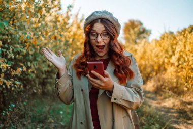 Sonbahar parkında ayakta durup cep telefonuna bakarken başarıyı kutlayan pozitif heyecanlı sonbahar kadınının fotoğrafı