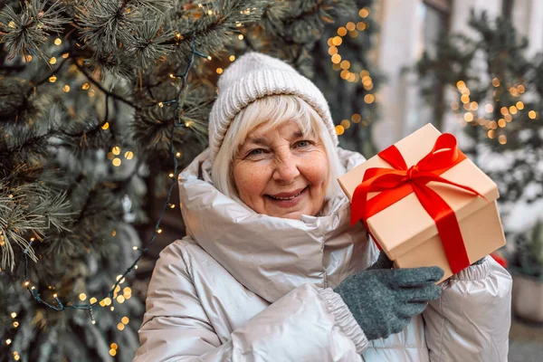 Merry Christmas and Happy Holidays. Senior woman having fun with surprise gift box present near Christmas tree outdoors at Christmas market on town hall square in Krakow, Poland. love, winter holidays
