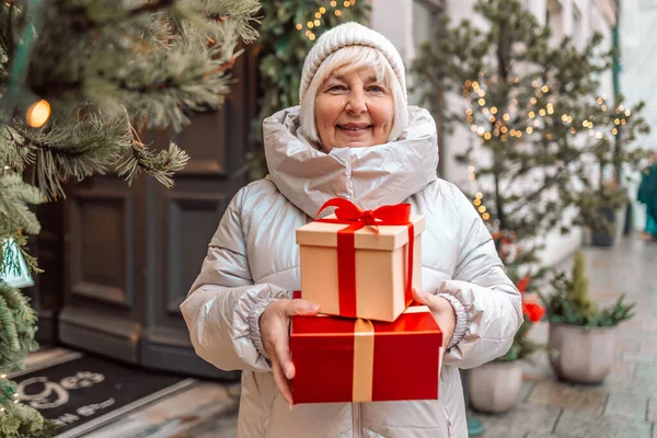 Merry Christmas and Happy Holidays. Senior woman having fun with surprise gift box present near Christmas tree outdoors at Christmas market on town hall square in Krakow, Poland. love, winter holidays