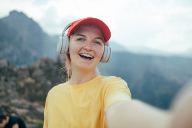 Kısa esmer saçlı, sarı bereli, sırtüstü sırtüstü sırtüstü gülümseyen genç bir kadın. Akıllı telefonuyla selfie çekiyor. Kayalık dağlarda zafer işareti yapıyor. seyahat, turizm, yürüyüş, teknoloji