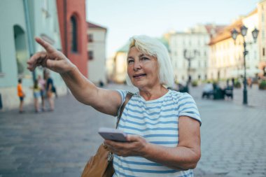 Bir el, şehirdeki bir sokağı gösteren boş ekranlı bir cep telefonu tutuyor. 50 'li yaşlarda, günlük giysiler içinde cep telefonu kullanan bir kadın, caddenin yönünü gösteriyor. 