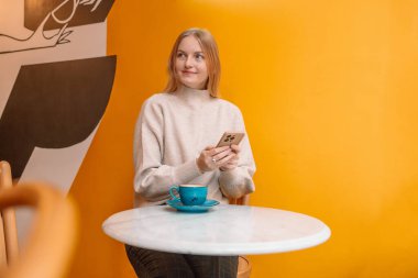 Çekici, genç, sarışın, gülümseyen bir kadın bir fincan kahveyle kafede sarı arka planda cep telefonu kullanıyor.