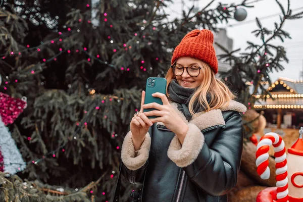 Happy blonde woman takes a picture of a European Christmas market with a smartphone. The girl enjoys the winter holidays season, visits the outdoor Christmas market, takes pictures on a mobile phone.