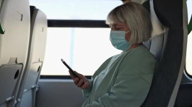 Senior woman in protective medical face mask in bus or train using mobile phone. First tourists, open boarders, new reality after covid 19. 
