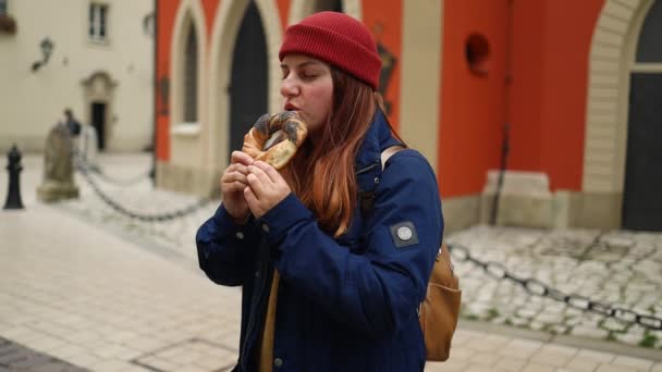 在克拉科夫旧城市场广场上 穿着鲜艳的帽子和秋季夹克的女游客拿着和吃烤奥布沃萨内克传统的美味佳肴百吉饼 优质Fullhd影片 — 图库视频影像
