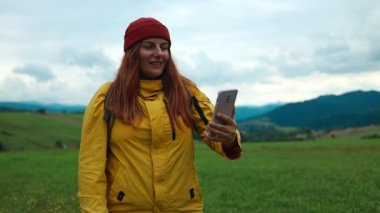 Video call. Happy traveler hiker girl in bright sportswear takes a selfie photo on a smartphone on top of a mountain on a summer day