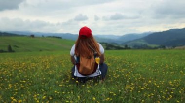 Tourist woman relax sitting in a grass field. Mountain range in background. Summer colorful scene. Backpack girl enjoy view 