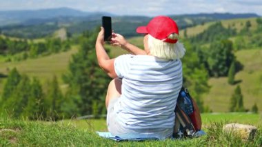 50 'li yaşlarda, sarı ceketli bir kadın selfie ya da dağ yürüyüşü fotoğrafı çekiyor. Uçurumun tepesinde kameraya gülümseyen mutlu yürüyüşçü barış jesti gösteriyor. Seyahat ve hobi kavramı. 