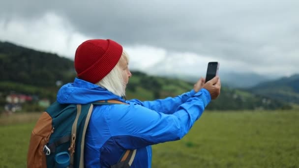 スタイリッシュな服のシニア女性ハイカーは 晴れた日に山の上に彼女の携帯電話のカメラでバックパッカーの旅行者のキャンパーをハイキング 美しい山の景色 ハイキング バックパッキング — ストック動画
