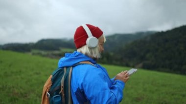 Back view of relaxed 50s woman listening music using wireless headphones among nature, enjoying scenic view of mountains, Poland. Girl use modern technologic device bluetooth earbud outdoor. 
