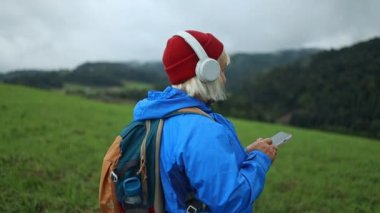 Back view of relaxed 50s woman listening music using wireless headphones among nature, enjoying scenic view of mountains, Poland. Girl use modern technologic device bluetooth earbud outdoor. 