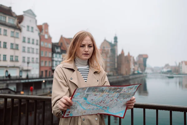 魅力的な若い女性観光客が新しい都市を探索しています ブロンドの女の子はグダニスクの街の通りに紙の地図を持ってる 秋のヨーロッパ旅行 — ストック写真