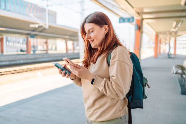 Seyahatin tadını çıkarın. Genç ve güzel bir bayan arka plandaki elektrikli trende akıllı telefon kullanarak istasyonda bekliyor. Turist mesajı ve demiryolu rotası, demiryolu konsepti. Yüksek