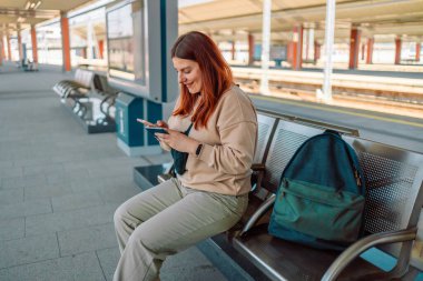 Seyahatin tadını çıkarın. Genç ve güzel bir bayan arka plandaki elektrikli trende akıllı telefon kullanarak istasyonda bekliyor. Turist mesajı ve demiryolu rotası, demiryolu konsepti. Yüksek