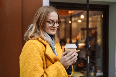 Parlak sarı palto giymiş hoş, mutlu genç bir bayan kahve tutuyor. Gözlüklü gülümseyen kız portresi Avrupa 'nın eski kasabasında çay ve kahvenin tadını çıkararak boş zaman geçirir.. 