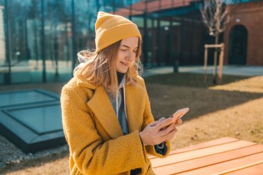 Genç gülümseyen mutlu öğrenci 30 'lu yaşlarda sarı ceketli ve şapkalı bir kadın. Şehir parkında bankta oturmuş dinleniyor. Cep telefonuyla internette sohbet ediyor.. 