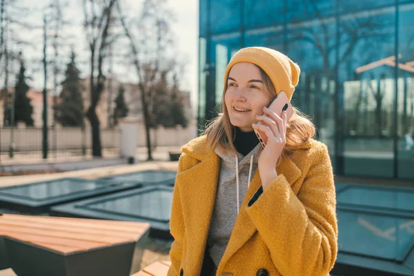 Stock image Horizontal shot of happy young woman makes telephone call wears casual sweatshirt and yellow coat had glad expression walks against blurred city building enjoys smartphone talking