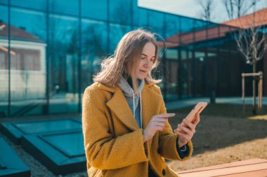 Genç gülümseyen mutlu öğrenci 30 'lu yaşlarda sarı ceketli ve şapkalı bir kadın. Şehir parkında bankta oturmuş dinleniyor. Cep telefonuyla internette sohbet ediyor.. 