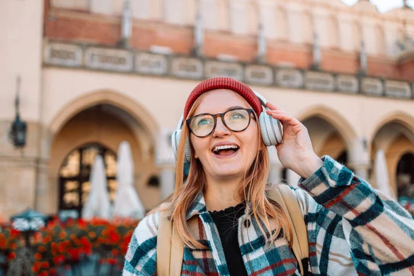 在欧洲克拉科夫市的大街上 年轻的金发女子用智能手机和耳机高兴地微笑着 高质量的照片 — 图库照片