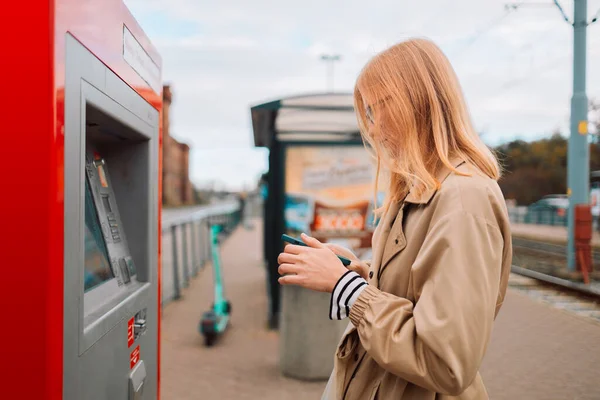 A side view of a young beautiful elegant blonde woman paying for service underground parking or buying a subway or train ticket using an electronic self-service kiosk