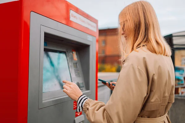 A side view of a young beautiful elegant blonde woman paying for service underground parking or buying a subway or train ticket using an electronic self-service kiosk