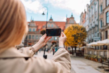 Şapkalı mutlu Avrupalı genç bayan turist Polonya 'nın eski Gdansk kentindeki akıllı telefondan fotoğraf ya da video çekiyor. Seyahat blogu, tatil yolculuğu, sosyal medya, aktif uygulamalı turizm