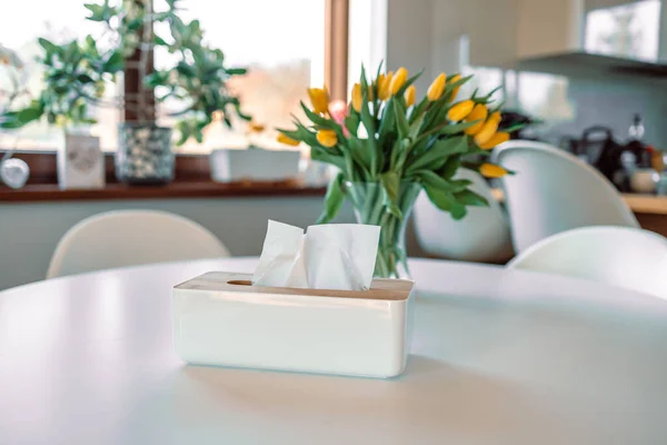 Tissue paper in napkin paper wooden white box on table. White sheet paper for cleaning face. Disposable cloth as hygienic. Spring flowers in glass vase on white wooden table in living room. Bouquet of