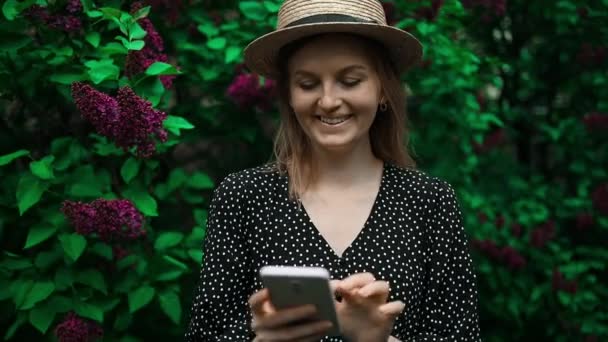 美しい若いブロンドの女性が自宅の緑の裏庭の庭の屋外で携帯電話を見ている 女の子はニュースを読み ソーシャルメディアでデータを共有し チャットし ミリ秒 — ストック動画