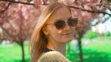 Tatlı sarışın kadın bahar çiçeğinin tadını çıkarıyor. Mutlu gülümseyen kız kameraya bakıyor. Temiz havada güneşli bir bahar. Şehir parkındaki bir sakura ağacının bahar çiçekleri. Süper yavaş çekim fotoğrafçılığı.
