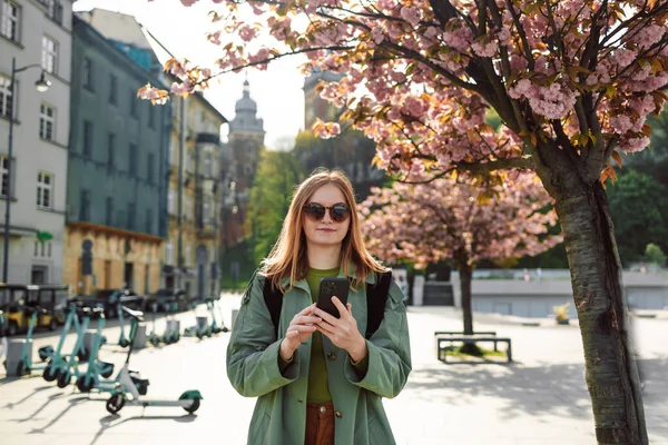 スタイリッシュな服ブロンドの女の子の観光客は ポーランドの春にWawelロイヤル城の近くにクラクフの旧市街を旅しながら スマートフォンの方向を探して旅します 春の桜 — ストック写真