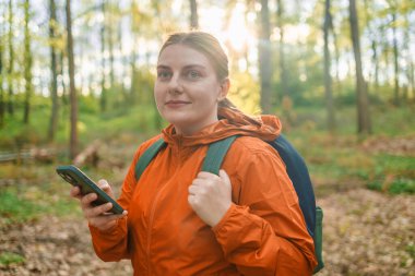 Sırt çantalı ve sıcak giysili genç bayan gezginin bahar ormanında telefon ekranına bakarken yan görüntüsü. Yürüyüş parkurundaki kadınlar. Ormanda tahta patika. Polonya 'daki Ulusal Park
