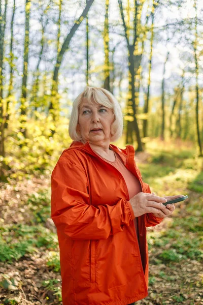 stock image Middle aged woman holding mobile phone in hands outdoors. Beautiful mature woman 50s in green sporty wear jogging in park and smiling, looking at her mobile phone app,