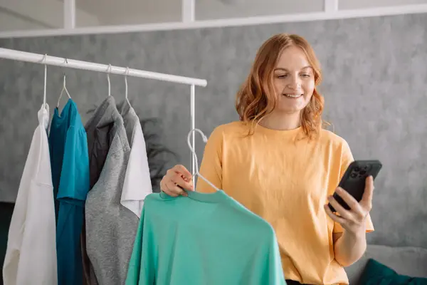 stock image Clothing, purchasing, style concept. Happy blonde haired woman chooses clothes at home wardrobe, holds comfortable jumper on hangers, outfits on racks in background, messages via mobile phone. High