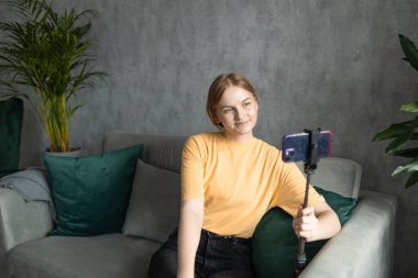 Sıradan giysiler içinde kadın blogcu koltukta oturup, tripoda yerleştirilmiş akıllı telefondan podcast 'i kaydetmeye hazırlanıyor.