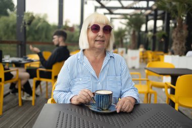 Happy 60s senior woman with stylish clothes and sunglasses drinking coffee at street cafe. Elderly, old and female enjoying an espresso on retirement feeling calm and confident. High quality photo