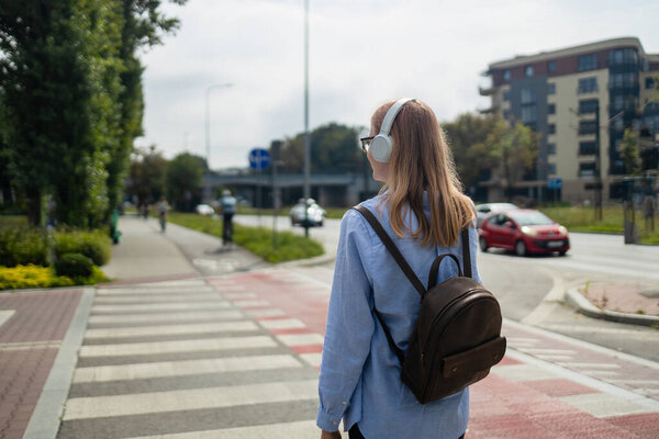 Back view of young woman with headphones woman listening the music, crossing street at traffic lights. High quality photo