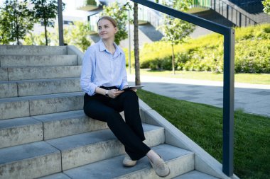 Portrait of successful professional businesswoman in smart casual using digital tablet outdoors in big city on busy street, smiling lady using smart business software for online work on pad computer