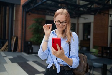 Neşeli, mutlu, şık bir kadın alışveriş merkezinin yanında dururken akıllı telefon uygulaması kullanıyor. Mavi tişörtlü mutlu kadın caddede yürürken akıllı telefonuna bakıyor.