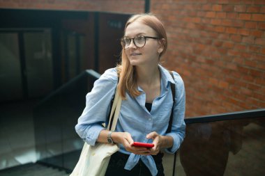 Neşeli, mutlu, şık bir kadın alışveriş merkezinin yanında dururken akıllı telefon uygulaması kullanıyor. Mavi tişörtlü mutlu kadın caddede yürürken akıllı telefonuna bakıyor.