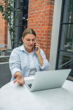 Üzgün, Kafkas asıllı, serbest çalışan 30 'lu yaşlarda bir kadın cep telefonuyla bilgisayar başında konuşuyor açık havada şehir kafesinde bulanık şehir binasına karşı oturuyor. Dizüstü bilgisayarda klavye kullanıyor. Bağımsız