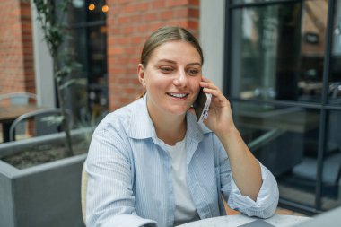 30 'lu yaşlarda serbest çalışan genç beyaz bir bayan cep telefonundan dizüstü bilgisayar kullanıyor açık havada şehir binasına karşı şehir kafesinde oturuyor. Dizüstü bilgisayarda klavye kullanıyor.