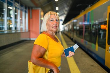 50 'lerin son sınıf öğrencisi kadın biyometrik pasaport ve tren istasyonunda tren bileti bekliyor. Hızlı trenle tatile git. Kaliteli fotoğraf.