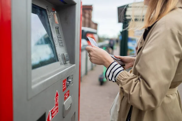 A side view of a young beautiful elegant blonde woman paying for service underground parking or buying a subway or train ticket using an electronic self-service kiosk. High quality photo