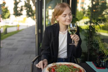 Genç iş kadını bir sokak kafesinde kameraya bakarak bir bardak şaraptan beyaz şarap içiyor. Şarap tatma. İtalyan şarabı. Arka planda bulanık ışık. Masada İtalyan pizzası. Boşluğu kopyala.