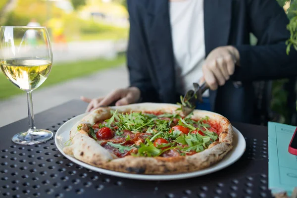 Kafkasyalı kadın müşterinin eli pizza keserken domates, prosciutto ve roka ile restoranın arka planında ya da kafede yuvarlak bir bıçak kullanması. İtalyan fast food. Kuru bir şarap kadehi