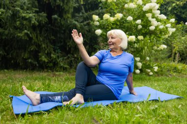Atletik 50 'lerin son sınıf öğrencisi kadın yoga minderinde poz veriyor. Spor kıyafetleri giymiş, parkta ya da ormanda tek başına. Yüksek kalite fotoğraf