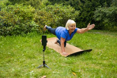 50 'li yaşlardaki kadın yoga minderinde pratik yapar, akıllı telefon kamerasında konuşur, takipçiler için video içeriği oluşturur, dışarıdaki tripodlarda cep telefonundaki webcam' e bakar. Spor Blog yazarı kavramı. 