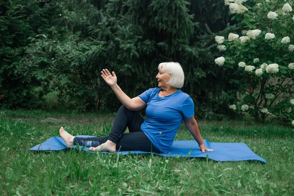 Athletic 50s senior woman doing poses at yoga mat outdoors dressed in sport clothes alone in the park or forest in nature. High quality photo