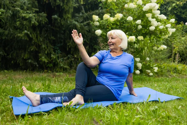 Athletic 50s senior woman doing poses at yoga mat outdoors dressed in sport clothes alone in the park or forest in nature. High quality photo